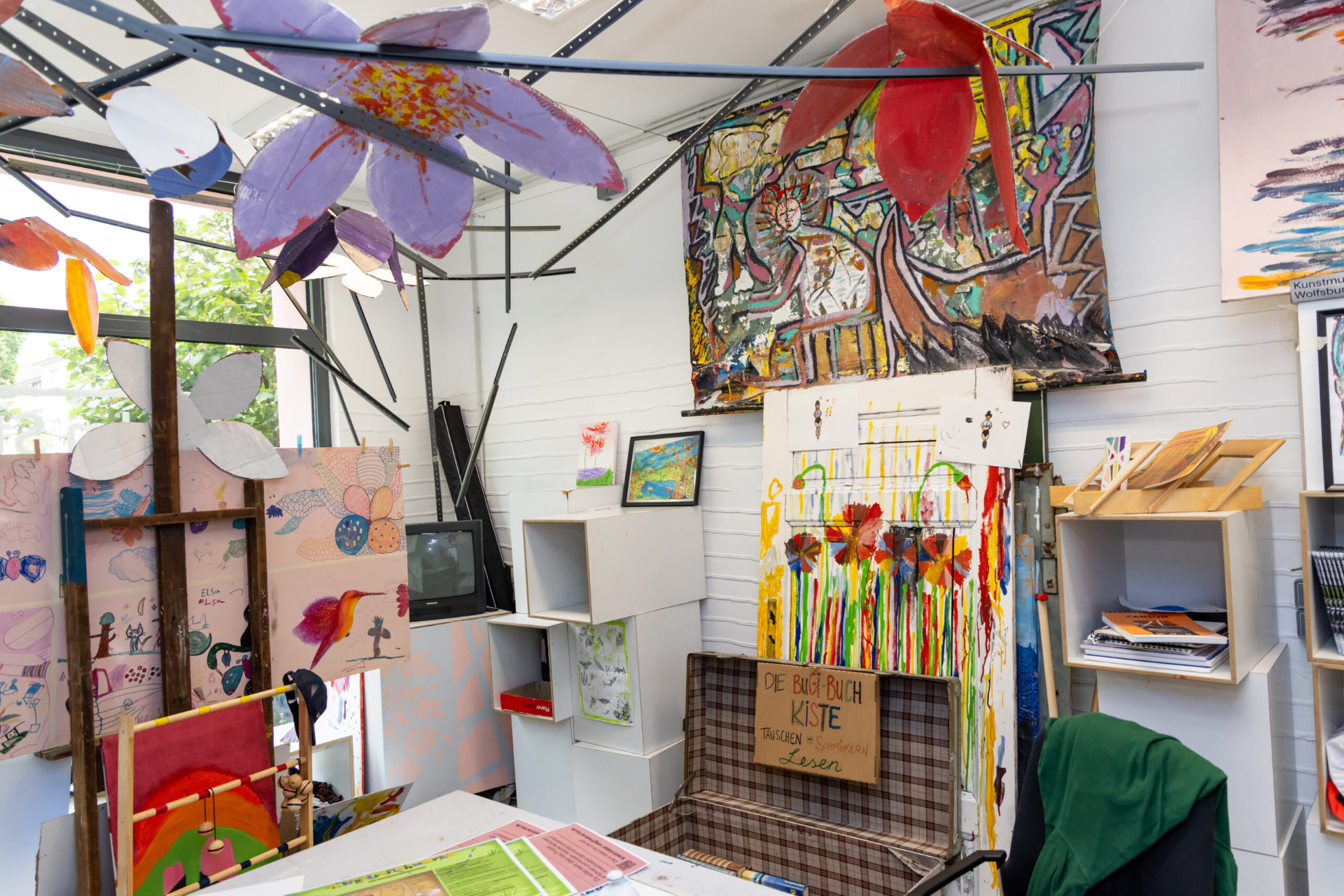 Atelierraum des Kreativlabors mit selbst gestalteten Blüten aus Karton, Gemälden von Kindern und einer Bücherkiste