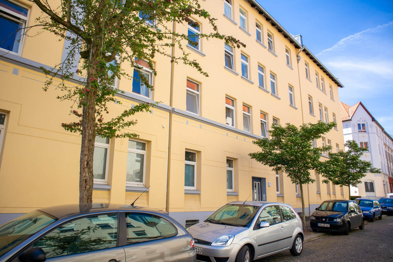Gelbe Altbaufassade mit hohen Fenstern, davor Bäume und parkende Autos.