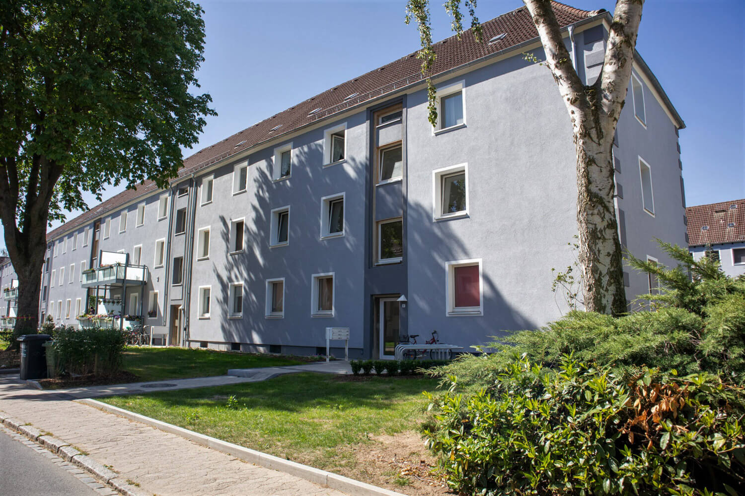Langgestrecktes graues Wohnhaus mit Balkonen, davor eine Rasenfläche mit Bäumen.