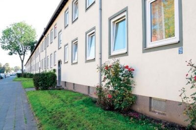 Wohnhauszeile mit heller Fassade, davor Rosenbeete und ein Gehweg.
