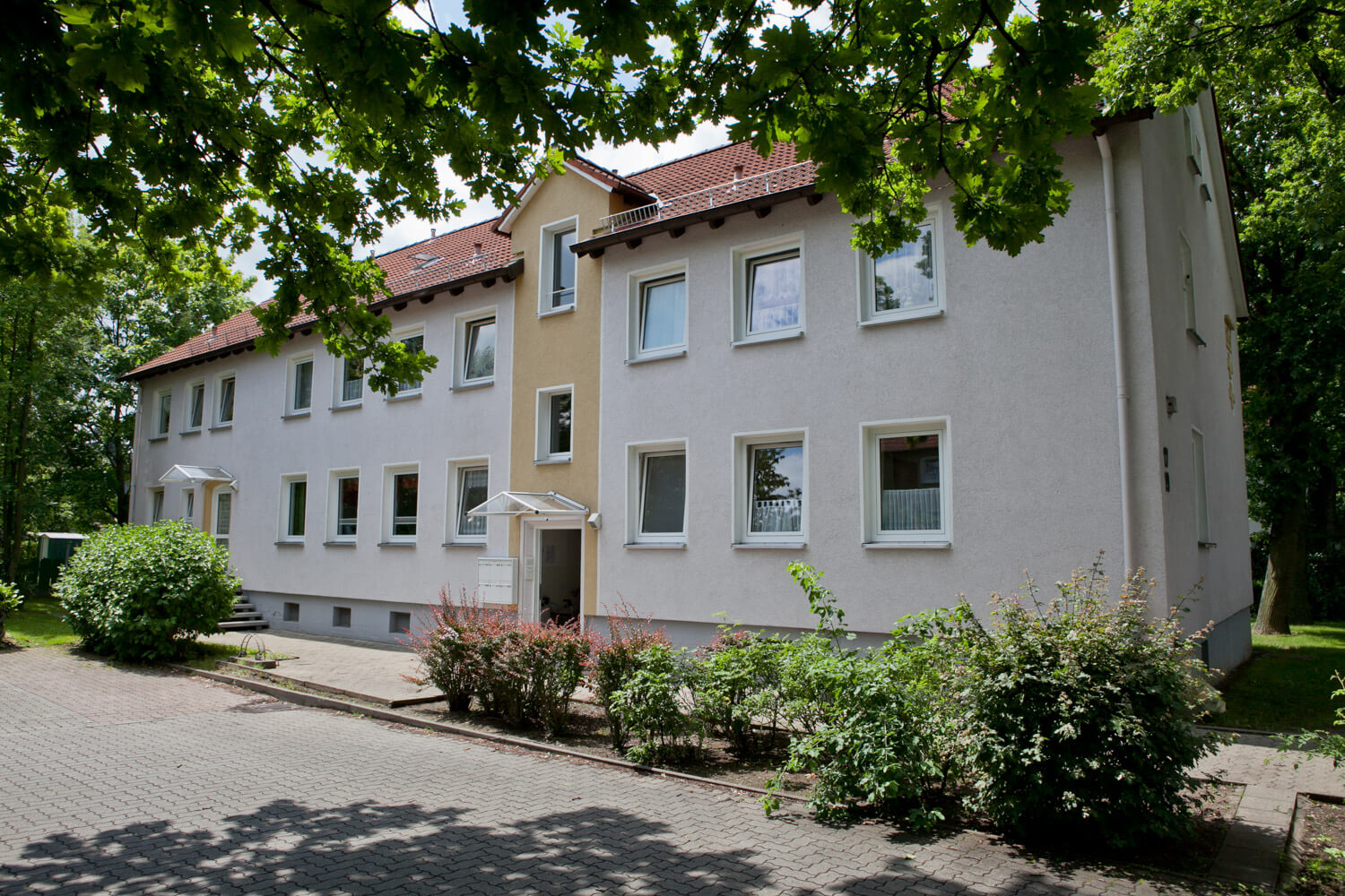 Helles Wohnhaus mit rotem Ziegeldach, umgeben von Bäumen und Sträuchern.